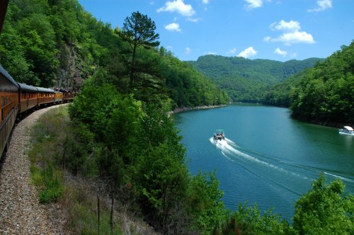 A scenic view of a river surrounded by lush green hills, with a train passing by and a boat on the water.