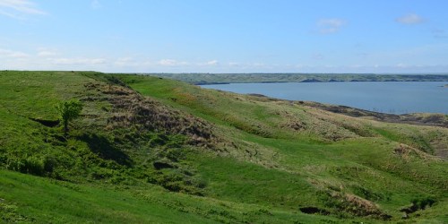 Lush green hills slope down to a calm blue lake under a clear sky, with distant hills visible on the horizon.
