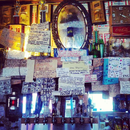 A cluttered bar wall covered with signs, notes, and memorabilia, featuring a mirror and various beer taps.