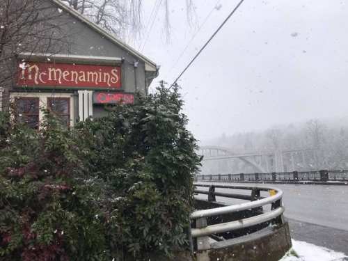 A cozy building with a sign reading "Open" amidst falling snow, with a bridge visible in the background.