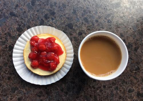 A cheesecake topped with cherry filling on a paper liner, next to a cup of brown coffee on a dark surface.