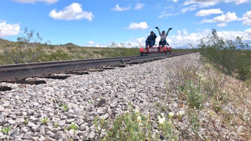 Two people joyfully riding a rail bike on a track surrounded by rocky terrain and sparse vegetation under a blue sky.