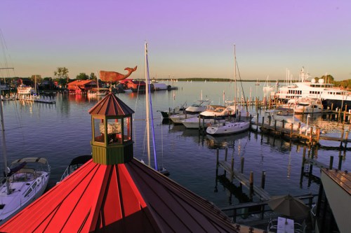 A scenic marina at sunset, featuring boats, a lighthouse, and a large whale sculpture in the background.