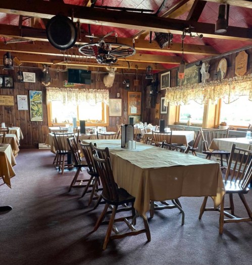 Cozy restaurant interior with wooden tables, chairs, and rustic decor, featuring large windows and warm lighting.