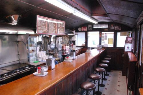 A vintage diner interior featuring a wooden counter, stools, and a menu board, with a grill and coffee station visible.