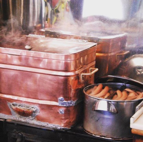 A copper steamer with a lid and a pot of sausages, surrounded by steam in a kitchen setting.