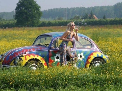 A woman in a colorful outfit blows bubbles next to a vibrant, painted vintage car in a field of flowers.