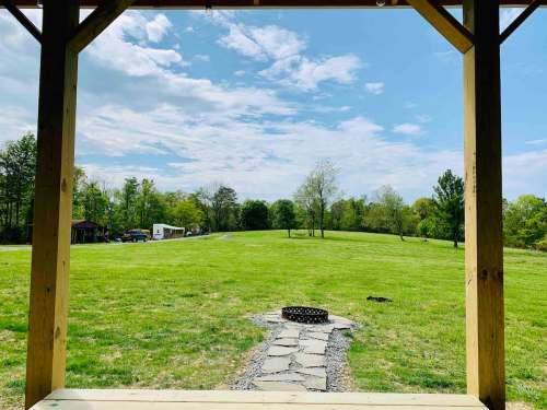 View from a porch showing a grassy field, trees, and a fire pit under a partly cloudy sky.