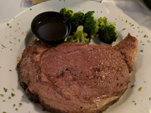 A plate featuring a juicy prime rib steak with a side of broccoli and a small cup of sauce.
