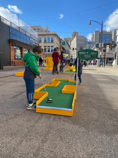 A child plays mini-golf on a street course with bright yellow obstacles and a green turf surface. City buildings in the background.