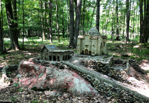 A miniature stone structure resembling a building, set in a wooded area with trees and greenery surrounding it.