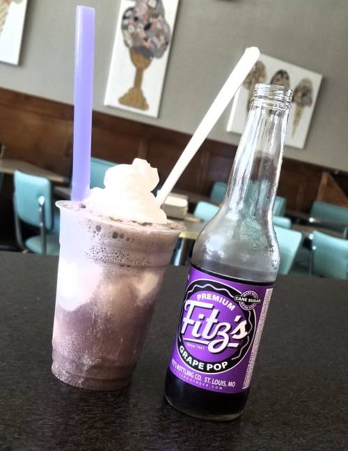 A frosty purple drink topped with whipped cream next to a bottle of Fitz's Grape Pop on a table.