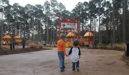 Two children walk hand in hand towards a colorful playground surrounded by tall trees.