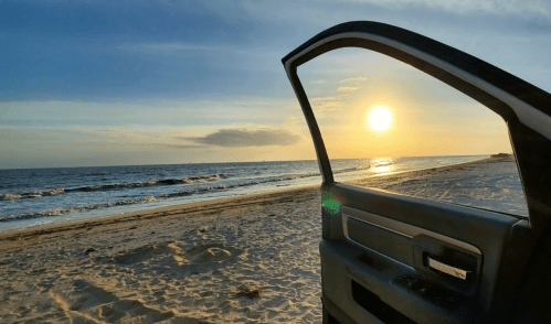 A beach at sunset viewed through an open car door, with waves gently lapping at the shore.