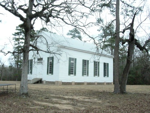 A white, historic building surrounded by bare trees, set in a grassy area with a cloudy sky above.