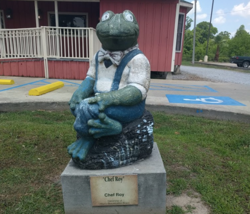 A frog statue named "Chef Roy" wearing a bow tie, sitting on a rock outside a building.