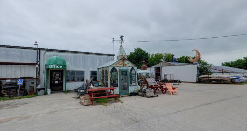 A rustic office building with a glass greenhouse, picnic tables, and a crescent moon sculpture in a cloudy setting.