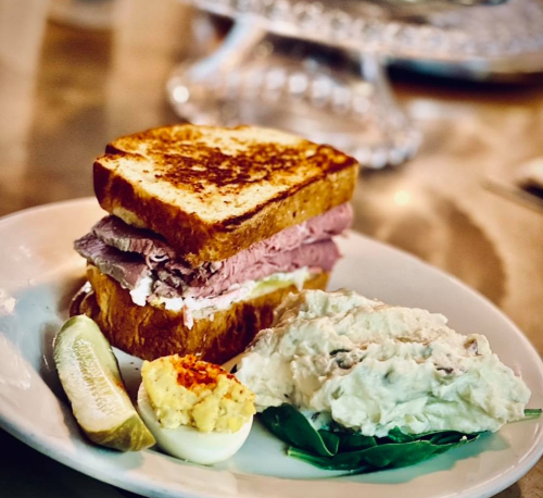A plate featuring a toasted sandwich with layers of meat, a slice of lemon, and a side of creamy potato salad.