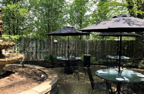 Outdoor dining area with black umbrellas, tables, and a fountain surrounded by greenery and a wooden fence.