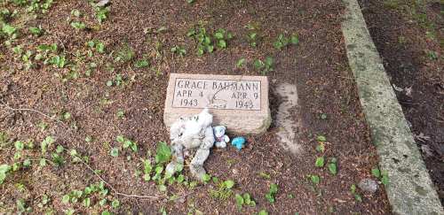 A gravestone for Grace Baumann, dated April 4, 1943 - April 9, 1943, surrounded by small toys and greenery.