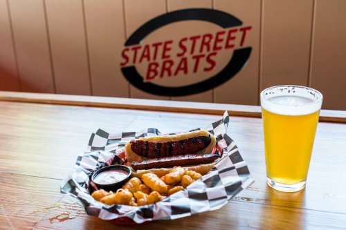 A plate with a grilled bratwurst, cheese curds, and a dipping sauce, alongside a glass of beer on a wooden table.