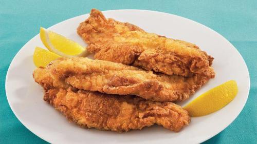 Three golden-brown fried fish fillets on a white plate, garnished with lemon wedges, on a teal background.