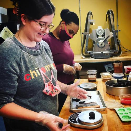 Two women in a kitchen, one weighing ingredients on a scale while the other prepares food, both wearing gloves.