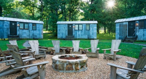 A cozy outdoor fire pit surrounded by wooden chairs, with rustic cabins in a green, wooded area.