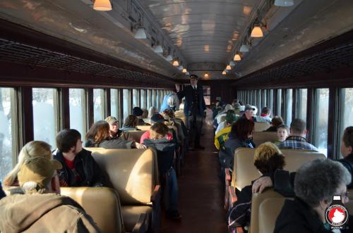 Interior of a vintage train car filled with passengers, some seated and others standing, with a conductor in uniform.