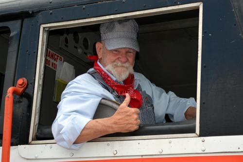 A smiling train conductor in a striped uniform and red bandana gives a thumbs-up from the window of a vintage train.