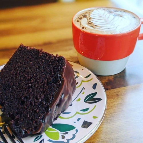 A slice of chocolate cake on a decorative plate next to a red and white cup of latte art on a wooden table.