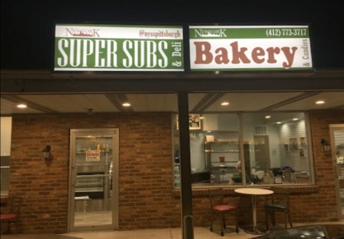 Signage for "New York K Super Subs & Deli" and "Bakery" on a brick storefront at night.