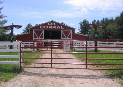 A red barn with "OK Corral" sign, surrounded by green grass and trees, with a gated entrance and a "No Parking" sign.