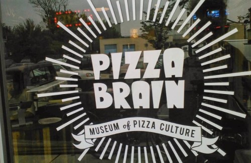 Sign for "Pizza Brain: Museum of Pizza Culture" with a sunburst design, displayed on a window.
