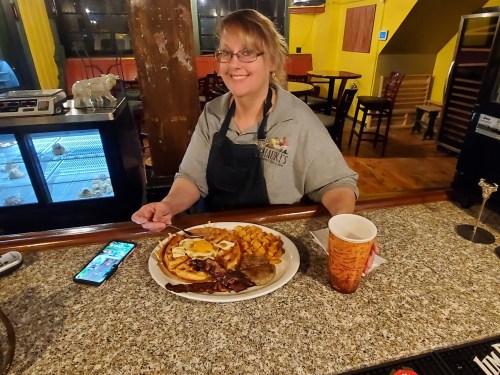 A smiling woman at a bar holds a plate of breakfast food and a drink, with a phone on the counter beside her.