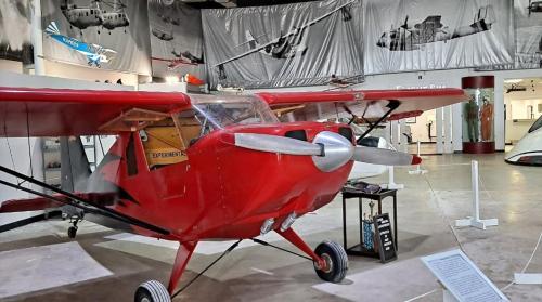 A red experimental airplane displayed in a museum, surrounded by vintage aircraft images and exhibits.