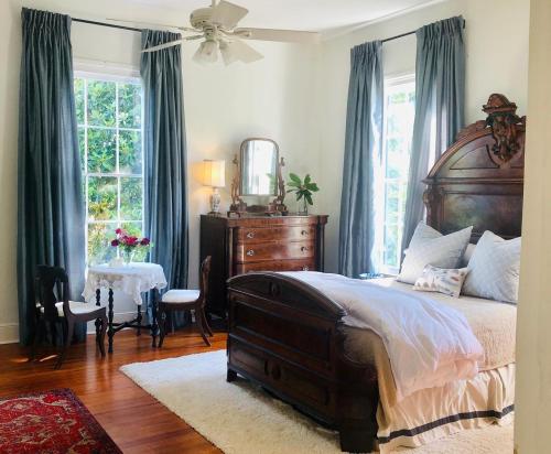 Cozy bedroom with a wooden bed, dresser, and a small table with flowers, featuring large windows and soft curtains.