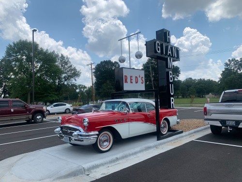 A classic red and white car parked in front of a diner sign that reads "Red's Giant Hamburgers" under a blue sky.