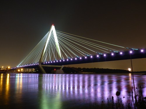 A brightly lit suspension bridge at night, reflecting colorful lights on the water below.