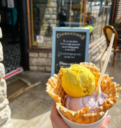 A hand holds a waffle cone filled with purple and yellow ice cream outside a charming shop.