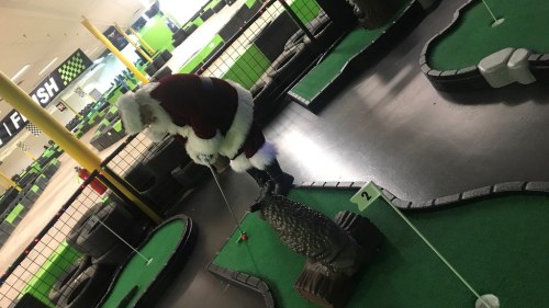 Santa Claus playing mini-golf on a colorful indoor course with green turf and obstacles.