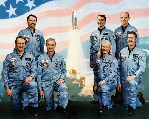 A group of seven NASA astronauts in blue flight suits, posed in front of an American flag and a space shuttle launch backdrop.