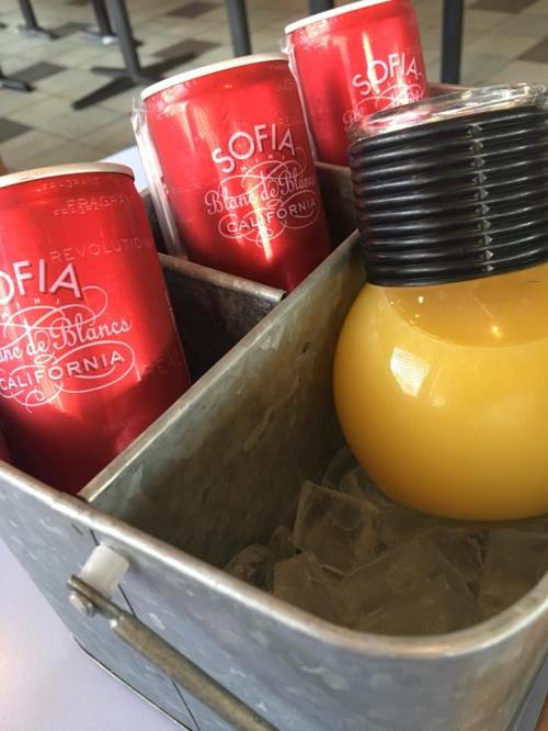 A metal bucket filled with ice, holding orange juice and three red cans of Sofia Blanc de Blancs sparkling wine.