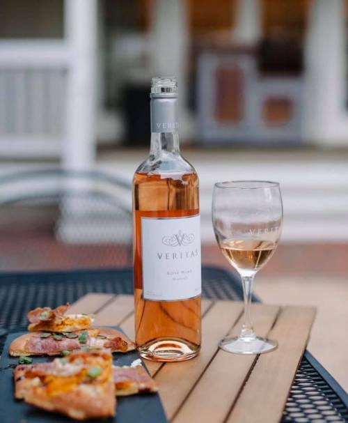 A bottle of Veritas rosé wine next to a glass and a plate of pizza on a table outdoors.