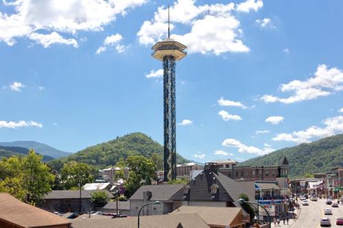 A tall observation tower stands against a blue sky with clouds, overlooking a town and green mountains in the background.