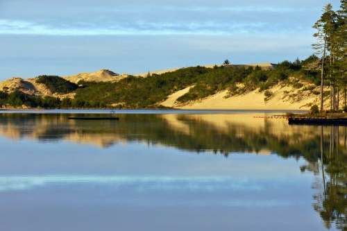 Serene lake reflecting sandy hills and trees under a clear sky, creating a peaceful natural landscape.