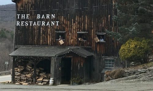 A rustic wooden barn with the sign "The Barn Restaurant," surrounded by trees and a gravel driveway.