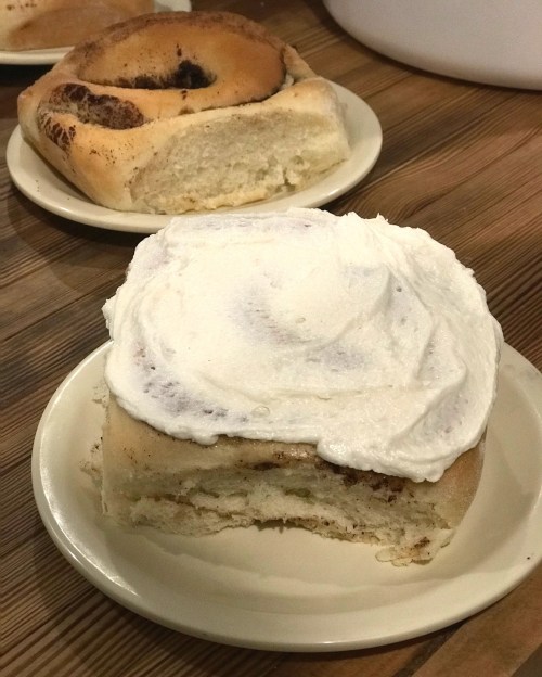 A frosted cinnamon roll on a plate, with a second roll in the background. Both are on a wooden table.