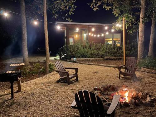 A cozy outdoor scene at night with a fire pit, string lights, and wooden chairs near a cabin surrounded by trees.