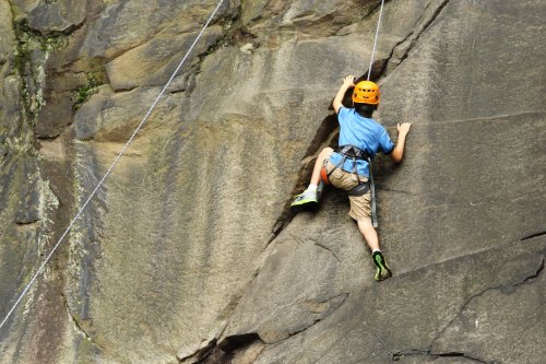 A child in a helmet climbs a rocky wall, using a harness and ropes for safety.